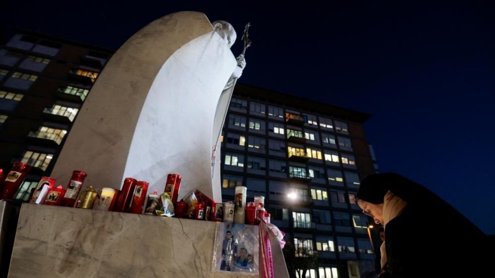 At night, a nun bows her head at the foot of a marble statue of the Pope, the base of which has been covered in candles.