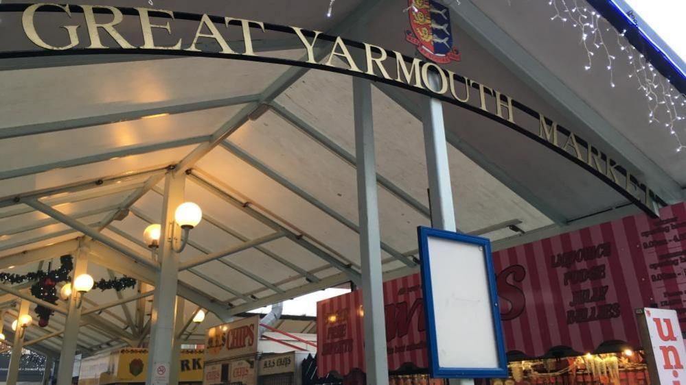 Great Yarmouth's old market building with the words Great Yarmouth Market cut out of metal with the borough crest above the arced text. Stalls are visible, as is the frame and canvas roof, and some lamps which are illuminated.