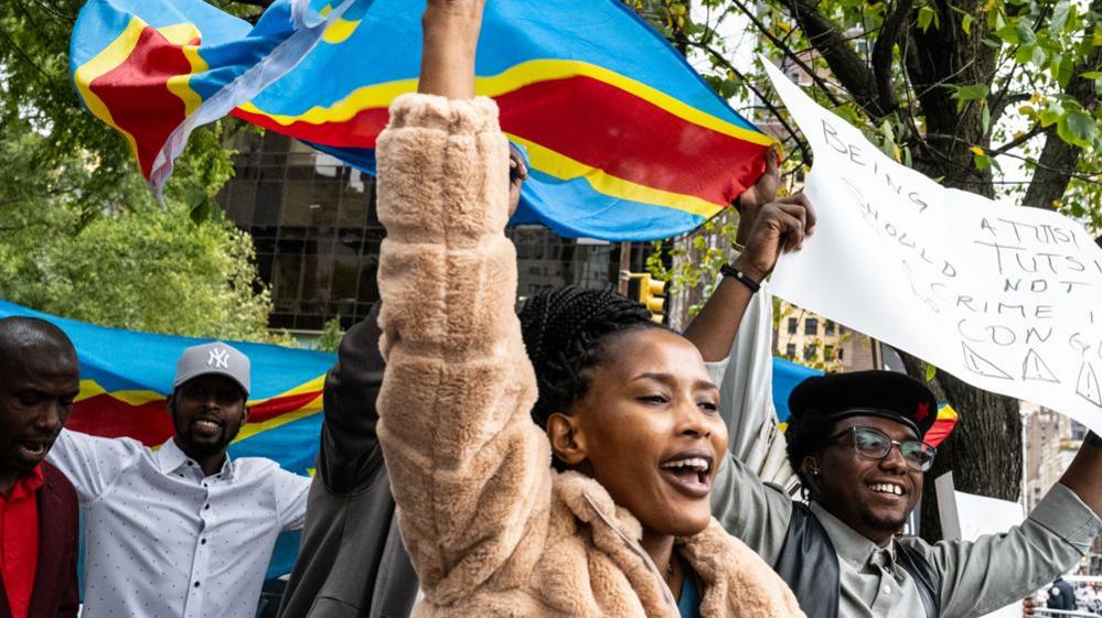 Protesters smile while raising Congolese flags and signs, one of which reads: "Being Tutsi should not be a crime in Congo"