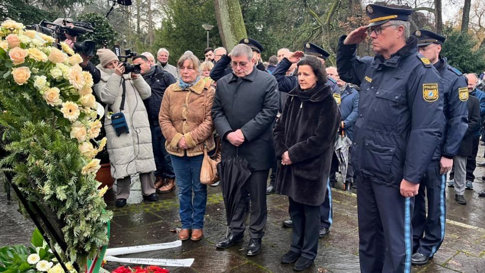 A police officer salutes and other people look on after a wreath of flowers is laid on a rainy day in a park in Bavaria where a toddler and a man were fatally attacked.