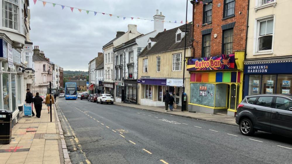 A bus drives up a newly opened road past two rows of terraced businesses and the demolition site. 