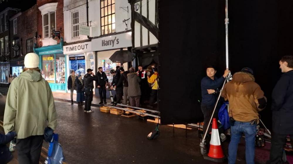 A dark street with a large black screen erected to one side, with a group of people gathered around camera equipment behind it.