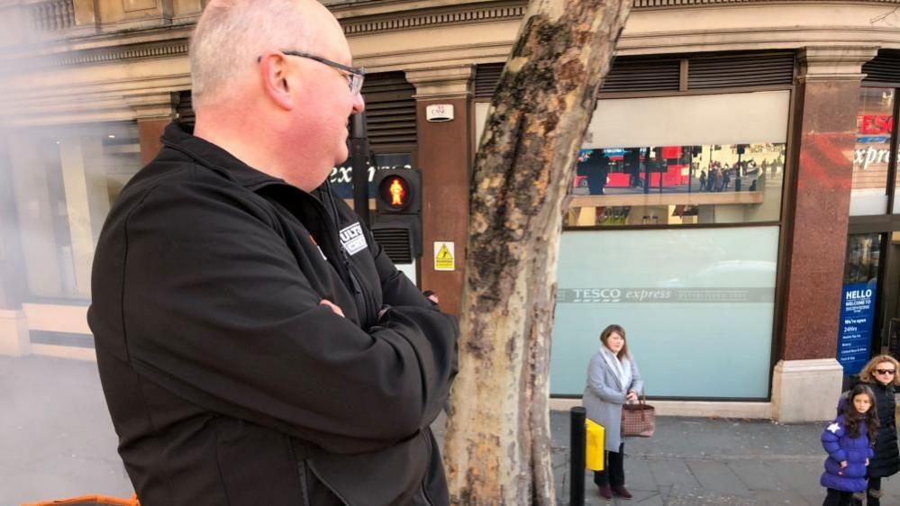 Mark Morrell with short white hair and glasses wearing a black T-shirt and looking down from the roof of an orange tank. People on the pavement below are looking up at him