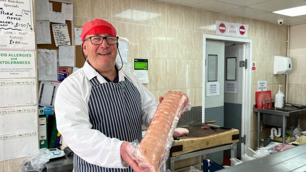 Jim Wyllie is wearing a butcher's white coat and blue and white striped apron, and a red flat cap. He is holding a joint of rolled stuffed pork belly. The back wall is tiled, and covered in notices and orders. There is a butcher's block and stainless steel sink and worktop.