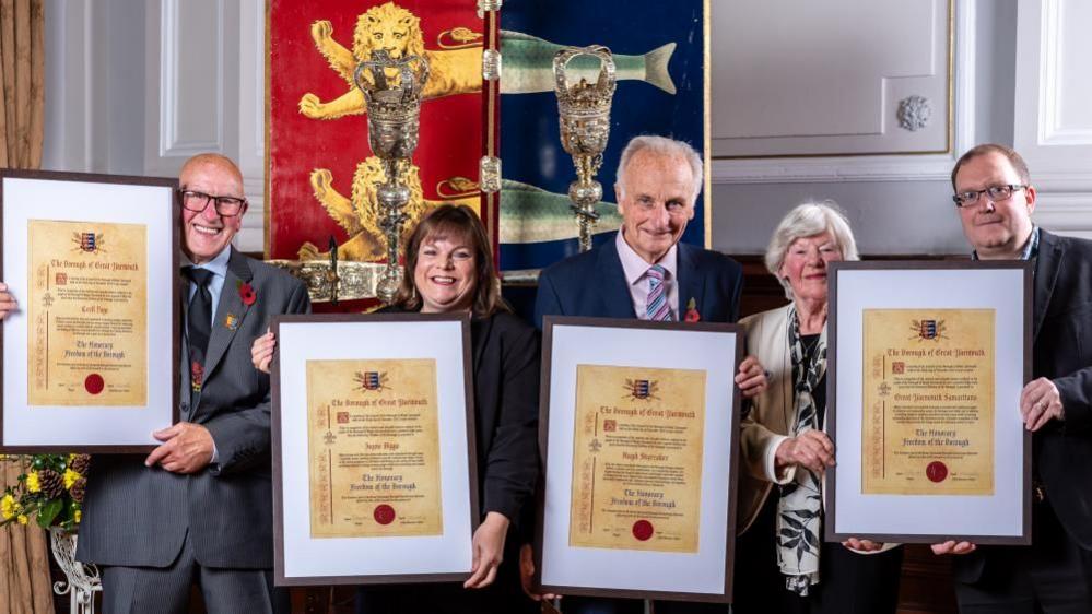 Recipients of the Freedom of the Borough stand from left - Cecil Page of the Kingfisher Boxing Club, Jayne Biggs of Norfolk Heart2Heart, Hugh Sturzaker of the Great Yarmouth Civic Society and members of the Great Yarmouth branch of The Samaritans. They are holding their scrolls of honour, mounted in frames, and stand before the staff and sceptre and the borough crest of Great Yarmouth. It is a shield painted red and blue divided vertically, with lion heads over the red segment, and herring fish tails over the blue, the heraldic image granted by King John in 1208.