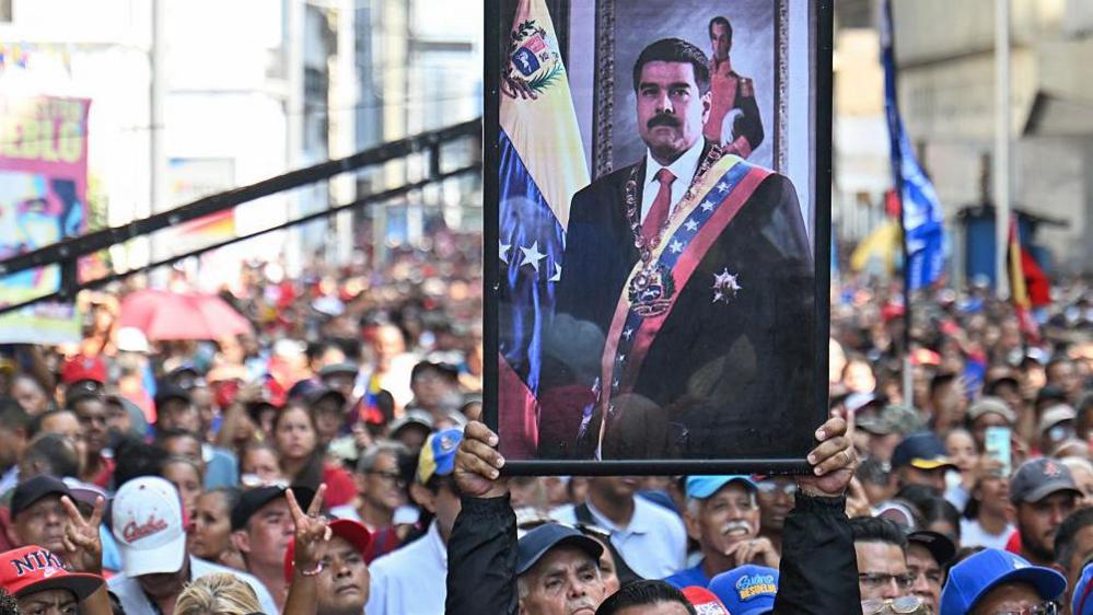 Një mbështetës i presidentit të rrëzuar të Venezuelës, Nicolás Maduro, mban portretin e tij gjatë një tubimi jashtë Asamblesë Kombëtare në Karakas të hënën. Qindra njerëz mund të shihen në turmë. Në portretin e Maduros, ai mban veshur një kostum të zi, kravatë të kuqe dhe një brez me ngjyrat e flamurit të Venezuelës.