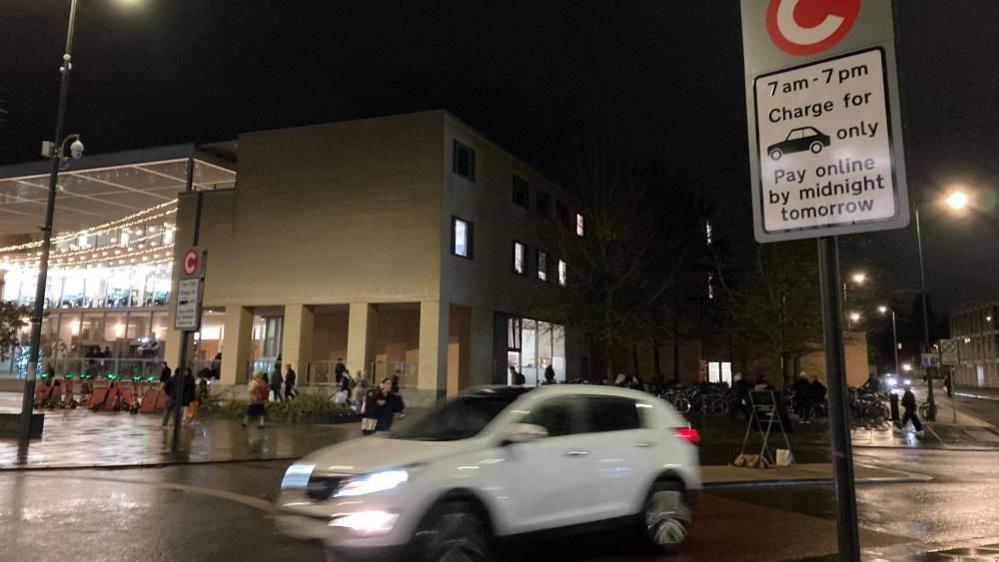 A white car is driving through a congestion charging point at night.