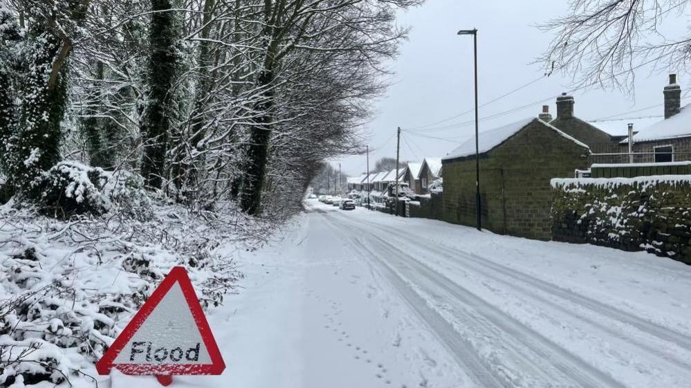 A picture taken in Sheffield show a road lined on one side by houses and on the other by trees. The scene is covered in snow and there are tyre tracks on the road. In the foreground is a sign warning of the possibility of floods. 