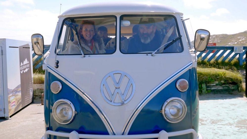 Jules and Gregg sit up front in a blue and white retro Volkswagen campervan on a sunny day. Their two sons, who are in their early twenties, sit in the back of the van.