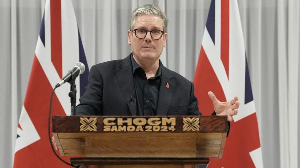 Prime Minister Sir Keir Starmer speaking at a podium which reads 'CHOGM Samoa 2024' in front of two Union flags