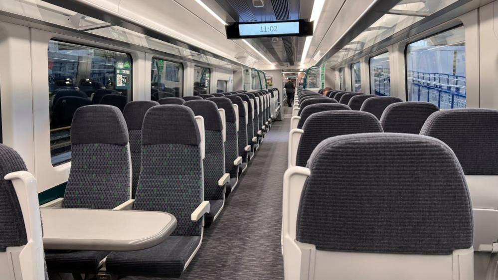 The inside of a new train, with lots of seats, with grey fabric and white tables. There is an aisle and rows of windows either side of the seats. A clock is in the middle the carriage saying the time 11:02.
