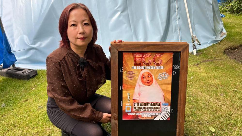 A person crouches on a grassy area beside a wooden sandwich-board sign displaying a colorful poster for EGG: The Biggest Unborn Artist. The person is wearing a brown long-sleeved blouse with a bow at the neck.
