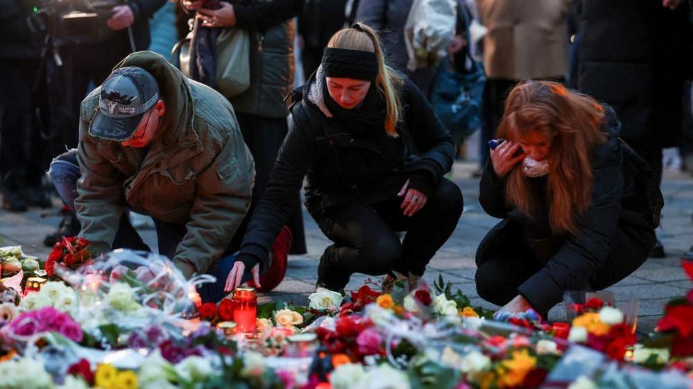 Three people - a man and two women - place tributes close to the scene of the attack. In the foreground can be seen flowers of various types and colours as well as candles in glass holders.