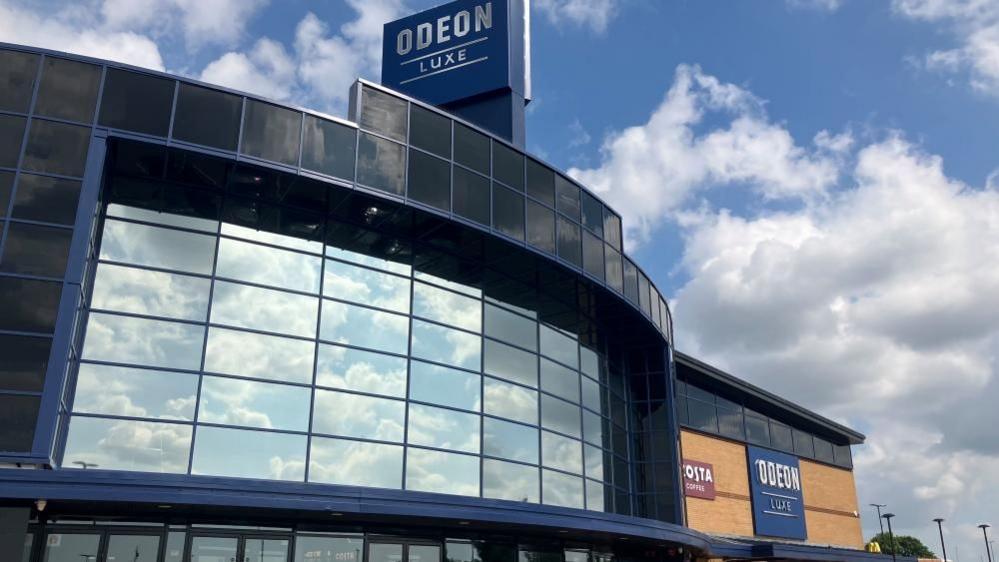 The outside of the cinema. It has curved reflective glass on the front and a sign above it