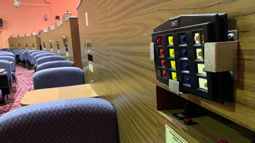 A row of seats to the left and to the right is a plastic bingo machine attached to a wood-panelled wall.