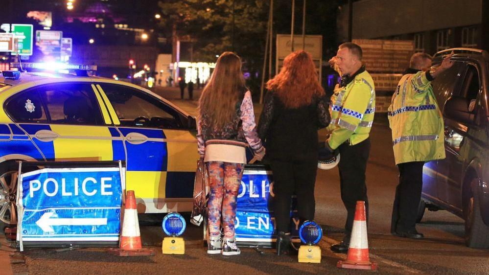 Two police officers stand in a road near Manchester Arena on the night of the bombing. One officer is speaking with a woman and a girl who are holding hands. The other officer is directing the driver of a car.