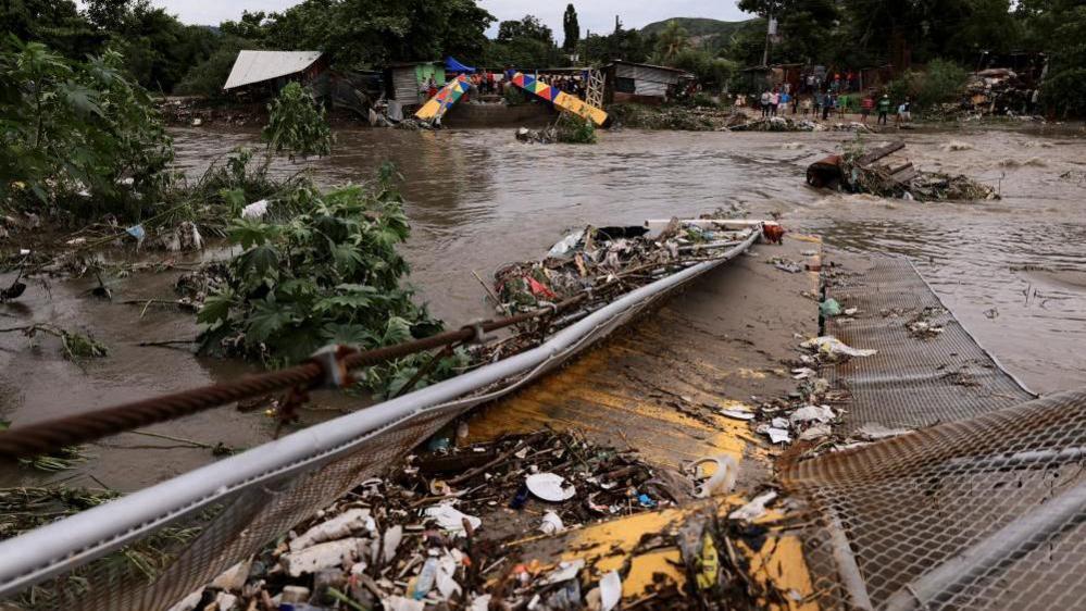 Twisted metal and debris can be seen partially submerged in the flooded waters of the Bermejo river. On the other side of the river's bank, people are standing and watching. 