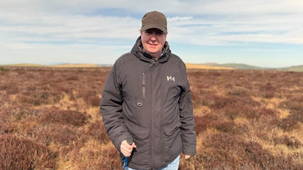 Prof Swindles is standing on a peat bog. He is smiling at the camera and is wearing a brown cap. He has a brown waterproof coat on with HH branding on the right lapel. He is holding a walking pole.