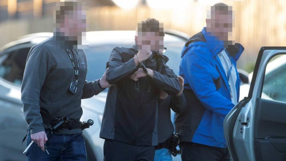 A handcuffed man is guided away by two police officers. All faces are blurred.