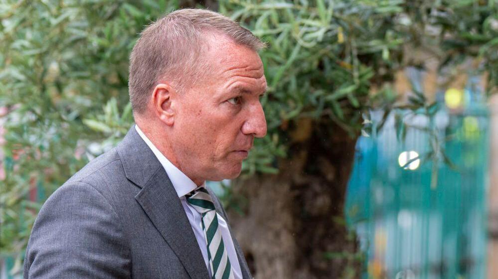 A man with short fair hair wearing a grey suit and green and silver tie looks off to the side