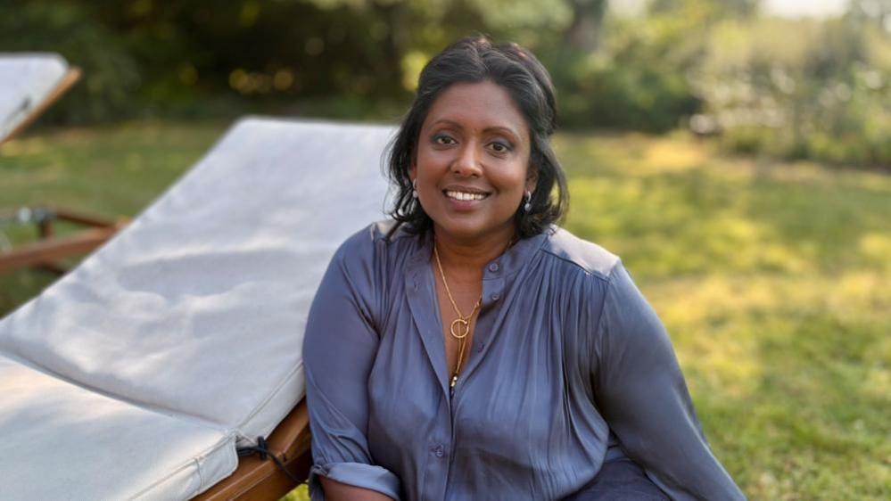 A Sri Lankan woman sits on the grass in a garden next to a white lounger. She has black hair and gold jewellery and is wearing a dark lilac silk shirt.