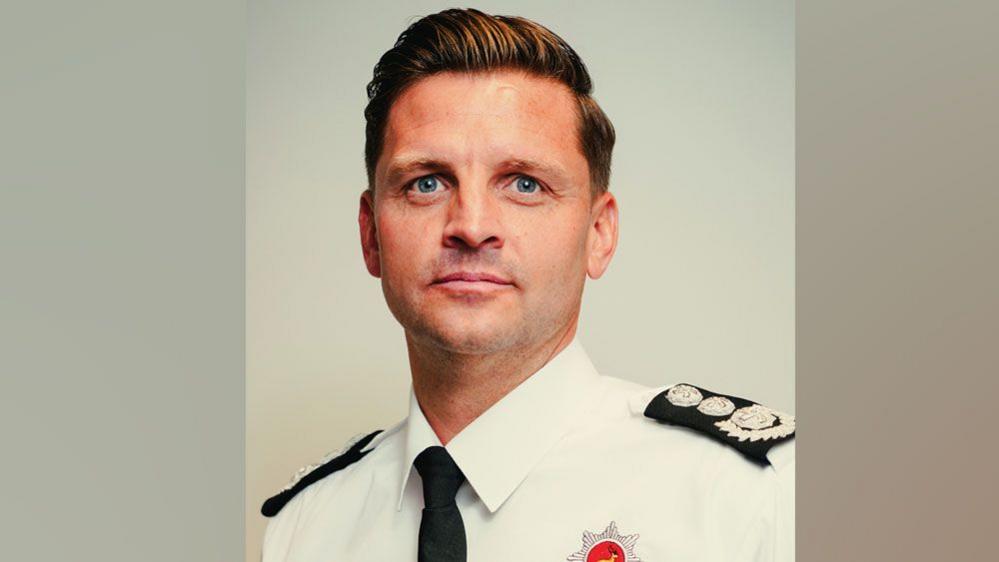 A man with gelled ginger hair swept sideways. He is in a smart white shirt with black tie and the top of a red fire service badge on his chest. 