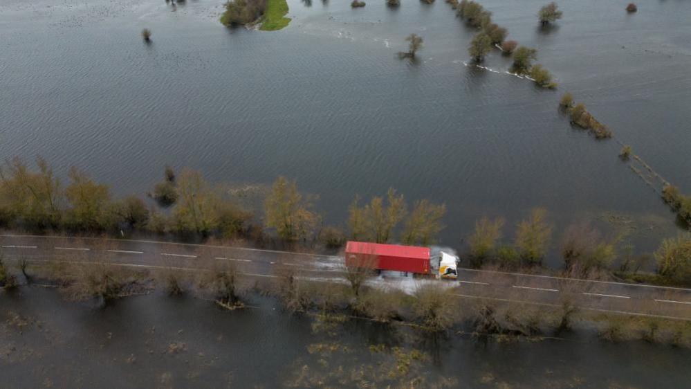 Red lorry drives through surface water on a road with flooded fields on both sides