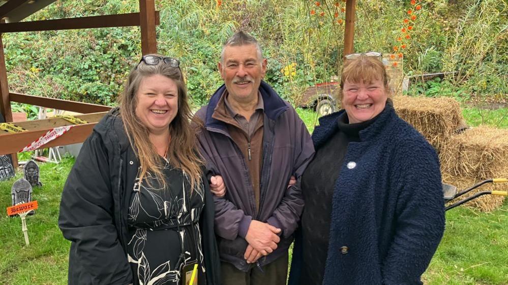 Three people: two women and one man pose on a farm - behind them are Halloween decorations and hay bales. The woman on the left has long brown hair with glasses on her head and is wearing a black floral dress. The man in the middle has short grey hair, a grey moustache and is wearing a short blue coat over a brown fleece jumper and the woman on the right has a beaming smile, is wearing a blue coat over a black jumper and has ginger hair.