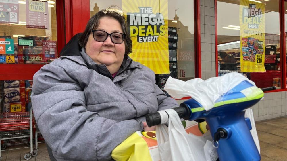 Sally Phipps is sitting in her mobility scooter outside Iceland, with bargains advertised in the windows. She has dark hair, has tinted spectacles and is wearing a grey jacket. She has a variety of carrier bags hanging from the handlebars of her scooter