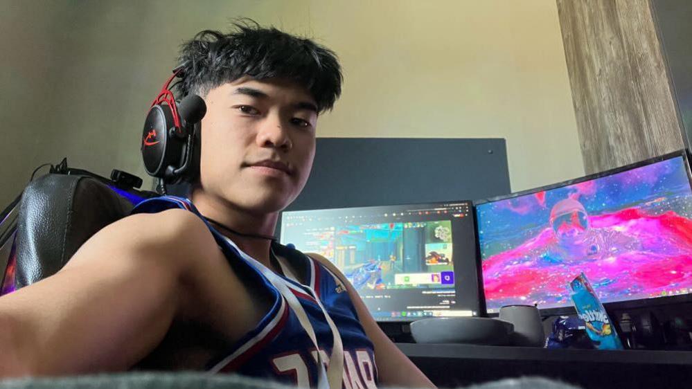 Dirk sits in front of his PC gaming set-up. He's wearing a headset and a basketball vest. Behind him are two monitors showing first-person shooting games. The desk has snacks, bowls and mugs on it.