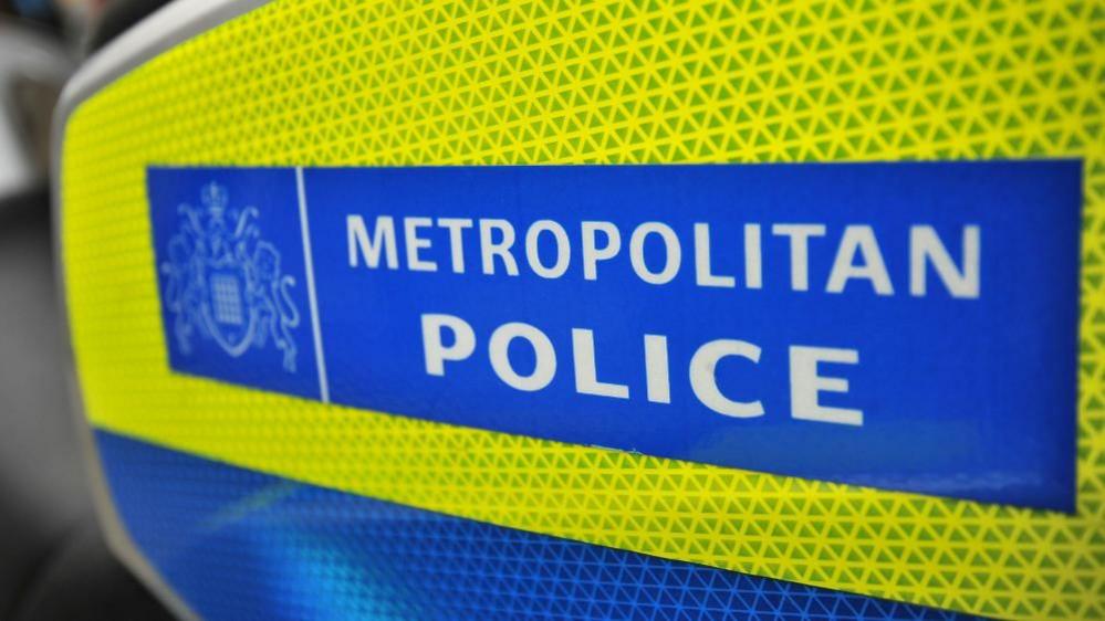 A sign on the side of a police motorcycle. There is a blue rectangular box with white writing reading 'Metropolitan Police' inside it, to the left of which there is another square box with the Metropolitan Police's coat of arms. This logo is on a yellow reflective background, with a blue reflective strip beneath it.
