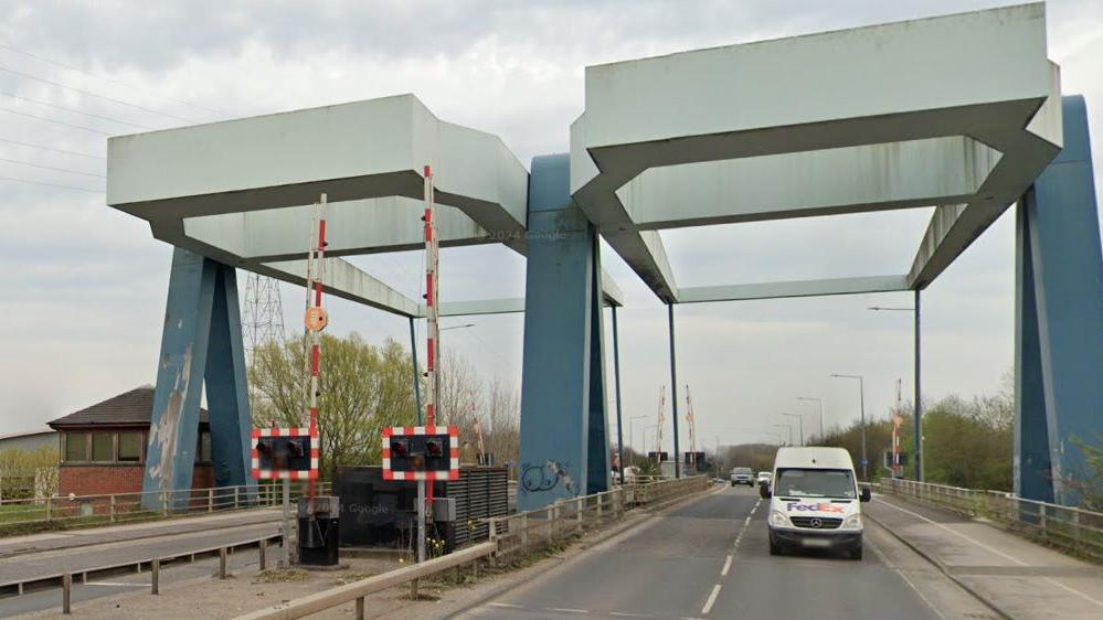 Twin blue and grey lifting bridges, which carry a dual carriageway over the River Hull. To the left are raised red and white barriers and warning lights, while to the right a white van travels towards the camera.