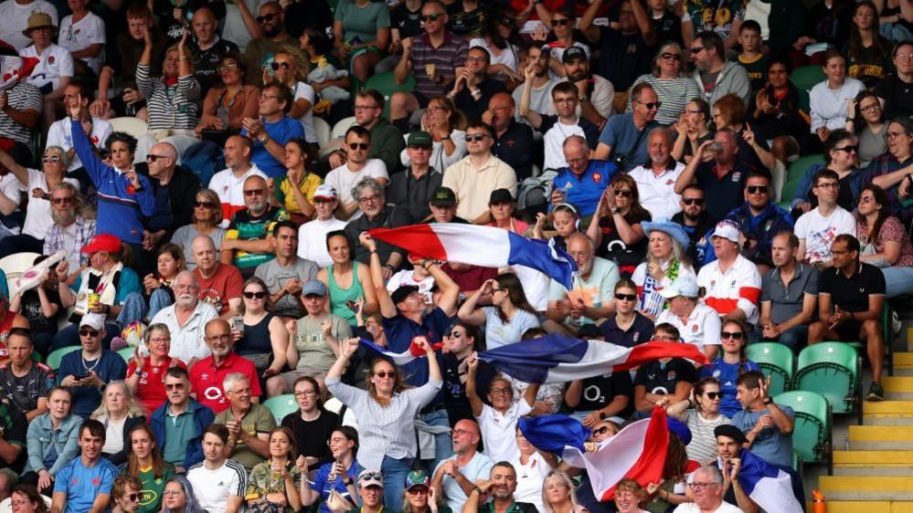 The crowd at Women's World Cup 2025 watching France v South Africa at Franklin's Gardens, Northampton. Many are holding flags, some are standing up but most are sitting down. 