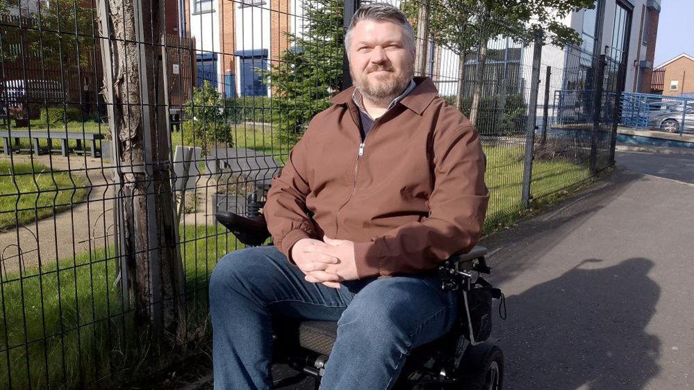 Johnny Harvey pictured on the pavement he has grey short hair and beard, he is in a wheelchair and wearing denim jeans and a brown zip up jacket. He is in front of barbed wire by a park and apartments.