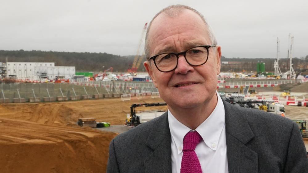 Patrick Vallance is  standing in front of a large construction site, with cranes and piles of dirt in the background. He is wearing a grey suit, white shirt and dark red tie. 