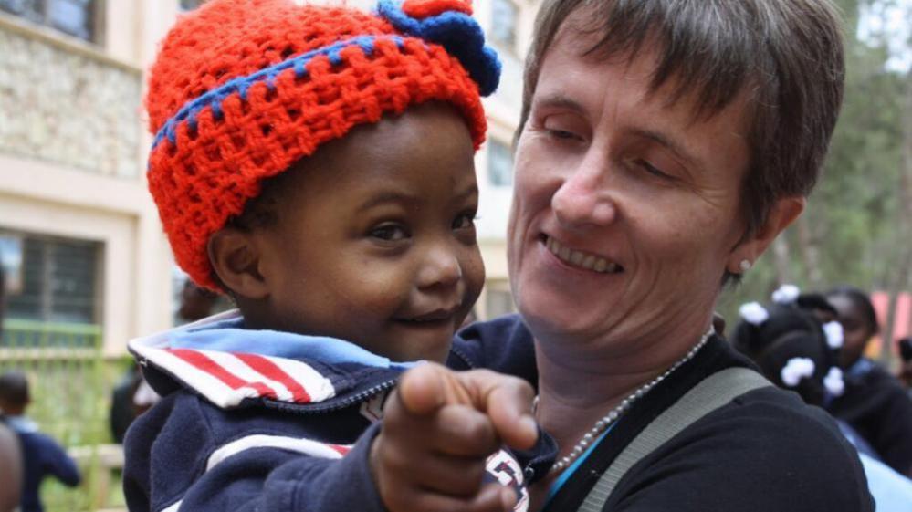Gena, right, is looking towards a child she is holding. She is smiling and has short brown hair. The child is wearing a red hat.