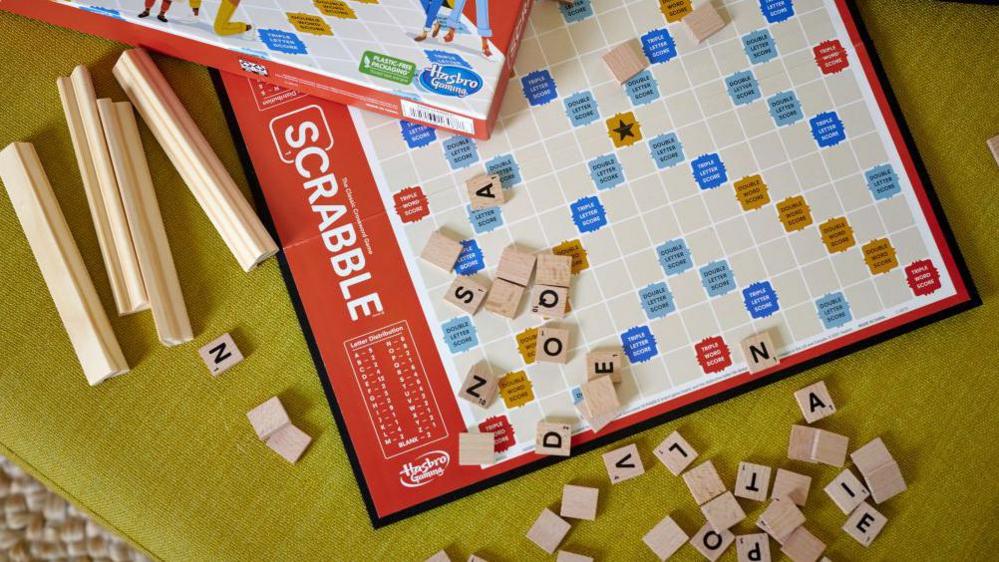 A Hasbro Scrabble board game arranged on a table alongside the box, tiles, and racks.