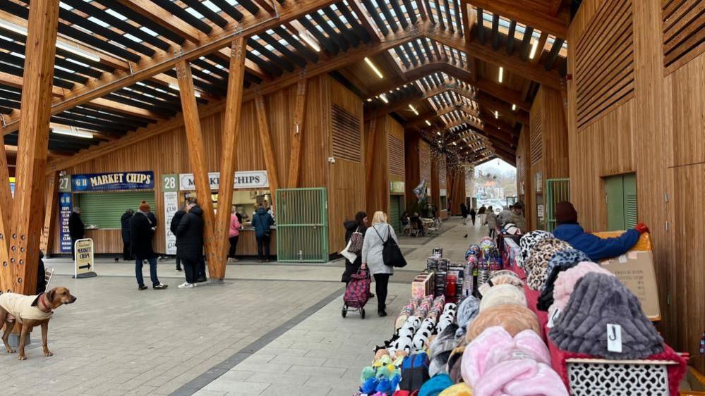 Great Yarmouth's market hall, a wooden structure with zinc roof. In the foreground is a market stall selling hats and toys. Beyond that are food stalls where a queue is formed. The picture shows the atrium of the market into the middle distance, with stalls either side, with louvred roof lights throughout.