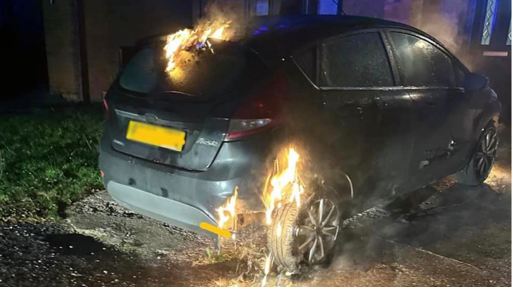 Flames around a rear tyre and top of a black car parked in darkness near a house in Rhosllanerchrugog