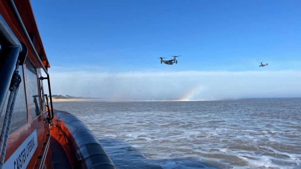 Two Osprey helicopters are pictured in the clear blue sky with a small rainbow created by the spray caused by the downdraught. The starboard side of the Caister Lifeboat all-weather vessel is visible on the left of the image with the coast and foreshore in the distant left.