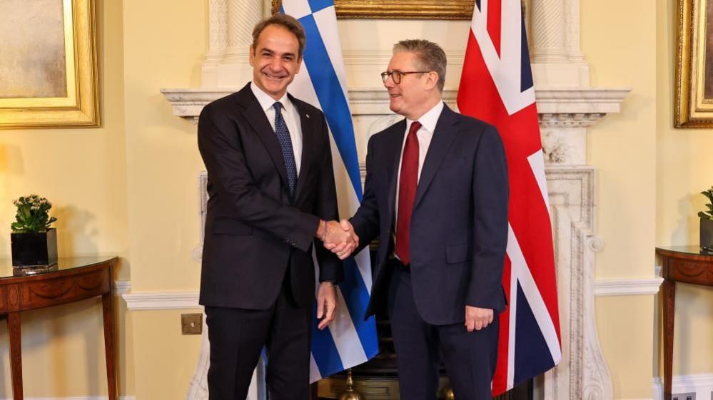 Mitsotakis, wearing a dark suit, stands in front of a Greek flag and shakes hands with Starmer, in a lighter suit, who is stood in front of a UK flag.