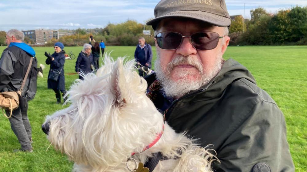 John Marriott with a white beard wearing a brown cap and coat carrying a white terrier on grass with some people behind him.