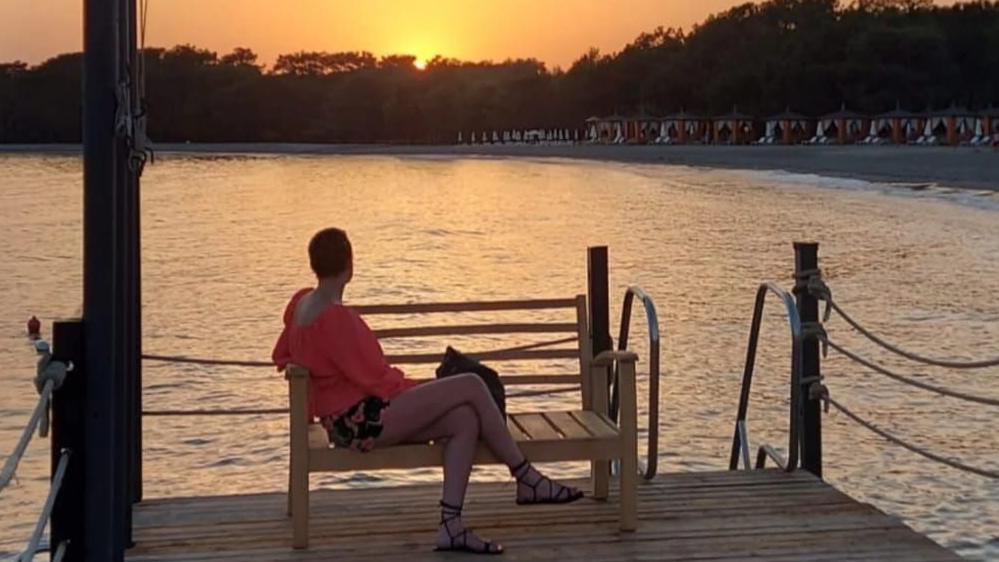 Rachel sits on a bench at the end of a dock looking out onto a bay with beach huts as a sun set in the back ground. 
She is looking out towards the water, has a loose pink top, shorts and sandals with her legs crossed. 