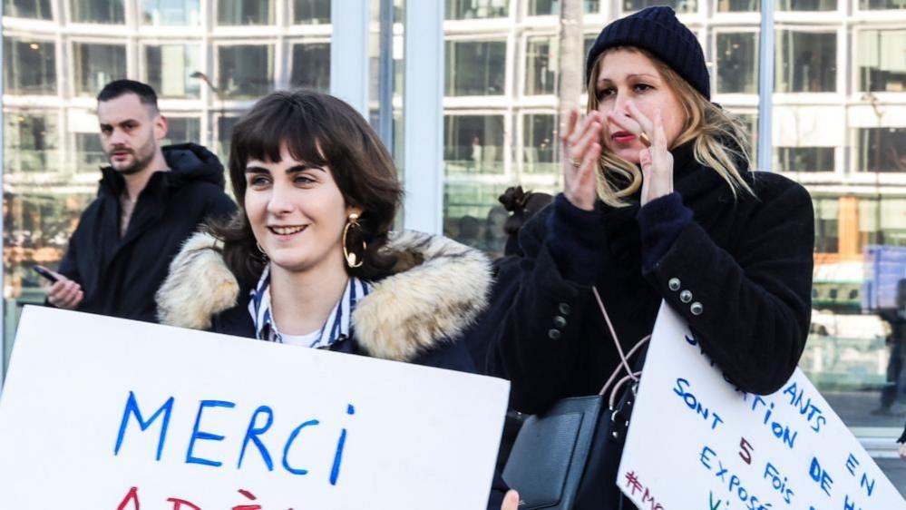 Two women holding signs written in French in support of the actor Adèle Haenel. 


The woman on the right is clapping and behind them a man looks on. 