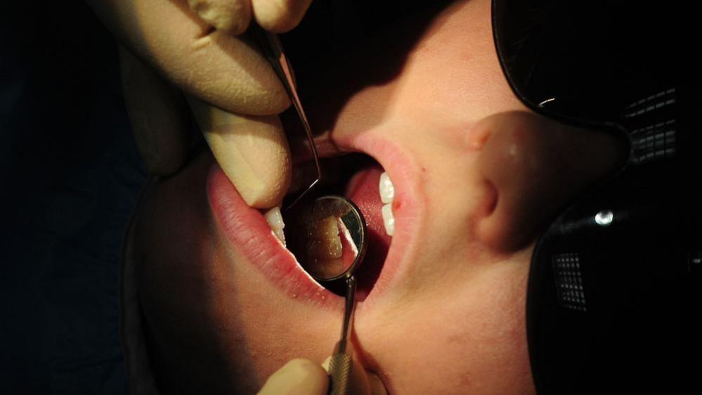 A dentist wearing latex gloves examines a patient's teeth, with the patient wearing protective glasses.