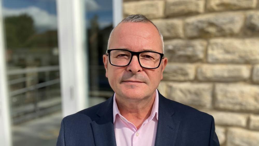 Lee Barron with short dark hair and glasses, wearing a dark blue jacket and pink shirt in front of a stone wall