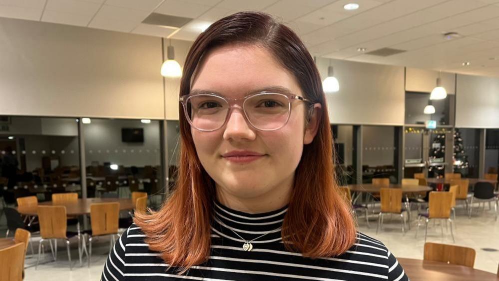 A young woman with black and red hair, wearing a stripy roll-neck top and glasses, stands in a rehearsal room with many wooden chairs and tables behind her. 