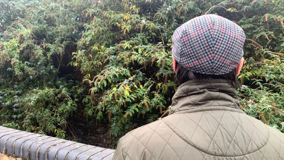 A man, 'Darren', stands with his back to the camera, he wears a green jacket and a flat cap. There are lots of tall bushes in the background.