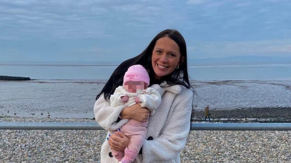 Kiena Dawes with long black hair smiling wearing a cream furry coat holding her baby dressed in a pink woolly hat, white furry coat and pink leggings and pink boots on the promenade with the sea in the background