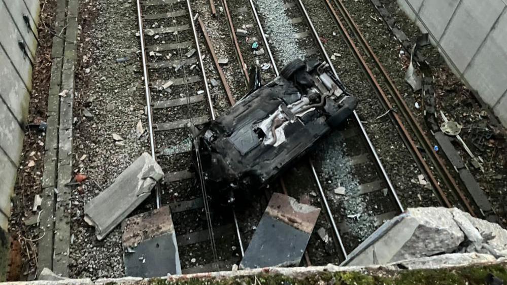 An upside down, crushed car lies on a railway line, with debris all around it.